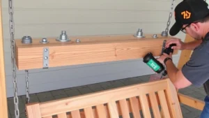 Carpenter assembling cedar porch swing frame with stainless steel bolts and galvanized chains attached to wooden beam structure, showing proper hardware installation and cross-bracing underneath seat platform.