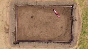 Aerial view of a properly graded and compacted soil base for a shed foundation, showing sloped ground directing water away, with string lines and level tools marking the perimeter