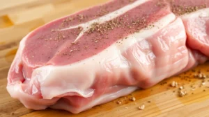 Close-up of raw pork loin with fat cap on wooden cutting board, seasoning visible on surface, natural lighting from above showing meat grain detail
