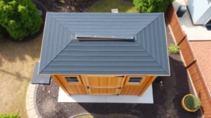 Aerial view of completed wooden garden shed with dark asphalt shingle roof, proper drainage gutters, and concrete foundation pads visible, surrounded by landscaping in suburban backyard
