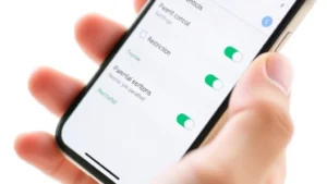 Close-up of a smartphone screen showing parental controls settings interface with toggle switches and restriction options enabled, modern iOS design aesthetic, hands holding phone against white background
