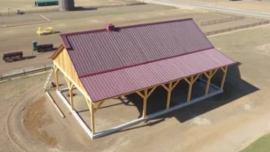 Aerial view of completed wooden pole barn with metal corrugated roofing and gable ends on rural property with cleared land, showing post spacing and structure foundation
