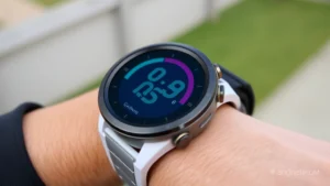 Close-up of a Garmin smartwatch screen displaying a colorful digital watch face with time, date, and heart rate metrics visible, worn on a wrist against a blurred outdoor background