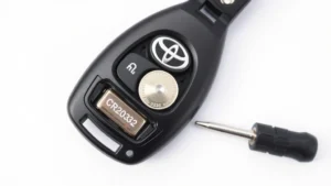 Close-up overhead view of Toyota key fob with battery compartment open, showing coin-cell CR2032 battery inside, flathead screwdriver beside it on white surface