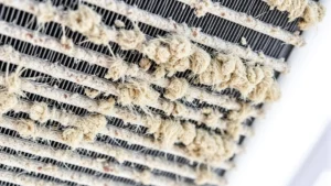 Close-up of refrigerator condenser coil fins covered in accumulated dust and pet hair, showing buildup on aluminum fins from above angle, natural lighting highlighting debris accumulation