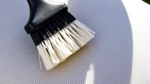 Close-up of soft-bristled brush gently cleaning textured outdoor cushion fabric with circular motions, showing dust particles being removed, natural daylight illuminating the work