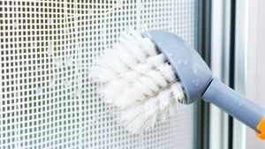 Close-up of soft-bristled brush gently scrubbing white fiberglass window screen mesh against aluminum frame, showing proper cleaning technique with soapy water droplets visible on mesh material